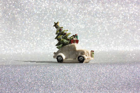 White toy truck car with green Christmas pine tree on roof rides on a silvered shining background. Minimalistic happy New Year's Eve background. Greeting card, copy space. Blue gift box delivery.の写真素材