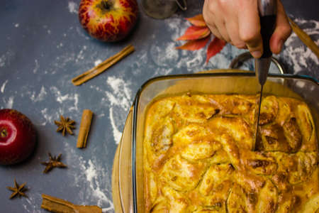 Woman holding a knife in hand, cutting a freshly baked apple pie in glass baking dish. Ingredients for baking - red apples, aniseed spices, cinnamon sticks, dusted flour top view. Thanksgiving dessertの写真素材