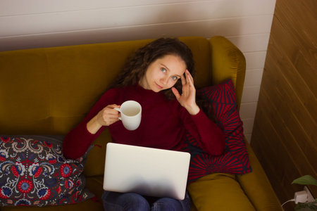 Pretty white young woman relaxing on couch at home looking at camera. Beautiful girl with cup of tea...