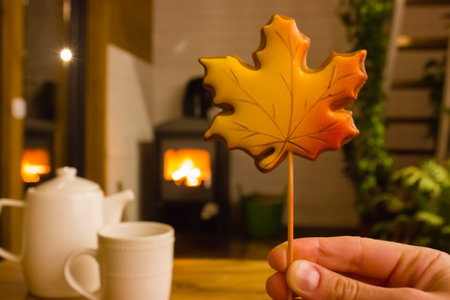 Maple-leaf-shaped maple leaf in woman's hand against blazing fire in fireplace background. Resting at cozy home at autumn evening. White teapot and cup stand on wooden table. Spend time in green spaceの写真素材