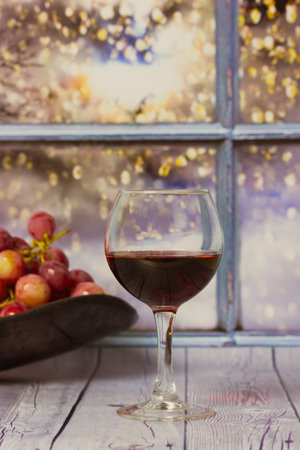 Bunch of purple grapes, glass of red wine stand on wooden table against window. Red grape on a plate. Harvest in vineyard. Romantic evening at home. Still Life with fruits and alcoholic beverage.の写真素材