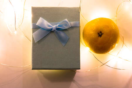 Blue gift box with a bow is lying on a table next to orange tangerine flatly. Glowing garland lights on white background. New Year celebration 2022 and Christmas, winter vacations. Present top view.の写真素材