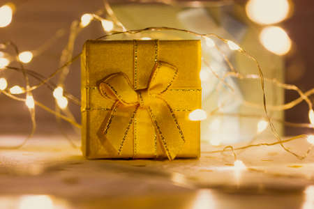 Yellow gift box with bow stands on table amidst glowing tangled led garland lights. New Year 2022, Christmas. Magical winter holidays shopping. Gold box with present in the evening. Festive backgroundの写真素材
