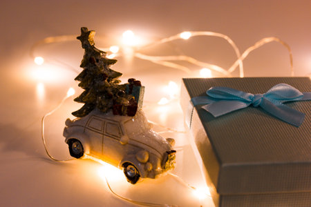 A small toy retro car with Christmas tree on a roof near a blue gift box with a bow stands amid tangled led garland with glowing lights. New Year's Eve 2022 concept. Winter shopping, sales, shopping.の写真素材
