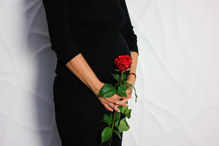 A pregnant woman in black dress is staying against a white wall with a red roseの写真素材