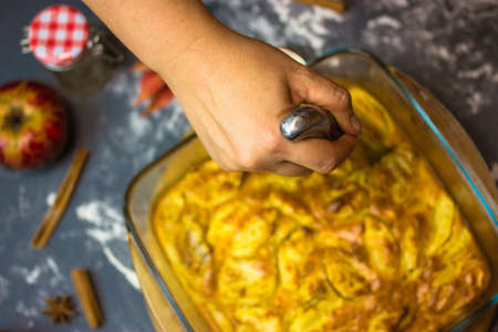 Woman holding a knife in hand, cutting a freshly baked apple pie in glass baking dish. Ingredients for baking - red apples, aniseed spices, cinnamon sticks, dusted flour top view. Thanksgiving dessertの写真素材