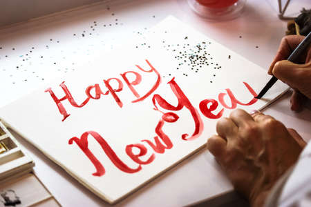 Artist draws DIY inscription Happy New Year with red paint, brush on white sheet of paper. Hand made postcard for Christmas 2022. Festive background, sequins in the form of stars. Painter creates art.の写真素材