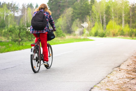 Kiev, Ukraine. May 1, 2020. A girl, young woman in checkered shirt, red pants, black backpack riding bicycle on a country paved road in woods, park in spring, summer. Journey alone, healthy lifestyle.のeditorial素材