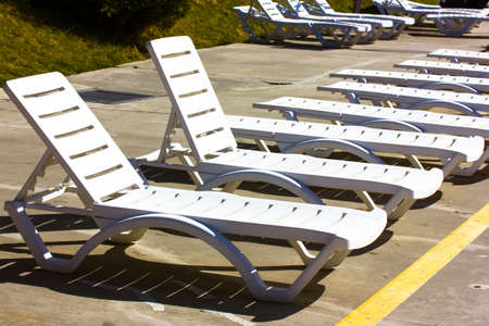 Empty white plastic sunbathing beds stand in a row on a beach or by the swimming pool at sunny day. Relaxation area, place to rest, chill out outdoor at summer vacations. Pool Furniture. Sun loungers.のeditorial素材
