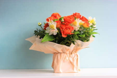 Beautiful bridal bouquet of different flowers alstromeria, small orange roses in cream colored wrapping paper stands on table on blue background. A gift for a woman, girl, mother, sister for March 8.の写真素材