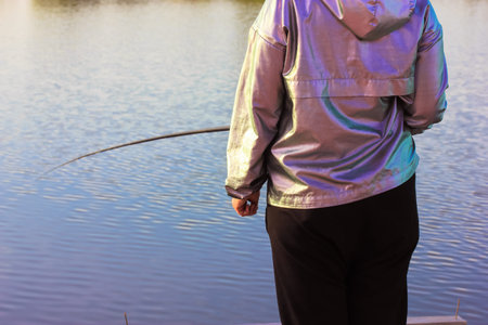 Fisherwoman with a yellow fishing rod by a pond, river, lake on a sunny spring day. Woman back view catching fish. Active leisure and recreation in nature reserve. Angling on a bank. bluewater.の写真素材