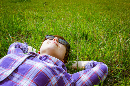 Smiling young woman in a plaid shirt, sunglasses lying relaxed on green grass with hands behind head viewed from above. Resting in a spring or summer sunny day on green lawn in park. earth day conceptの写真素材