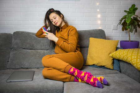 Stylish young brunette woman in warm brown tracksuit, colorful socks is lying relaxed on gray couch with smartphone in hand. A girl is using smartphone, gadget, online shopping, chatting. Cozy home.の写真素材