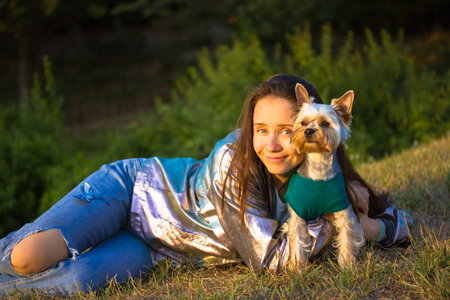 Beautiful brunette female, girl lying on lawn hugging beloved Yorkshire Terrier little dog....