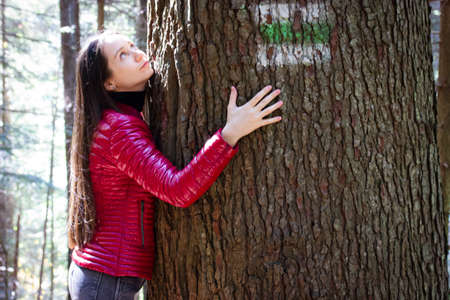 Beautiful white long-haired brunette young woman in red jacket hugging a huge tree trunk and looking up. Unity, oneness, feeling to become one with nature, recreation in woods, forest, national park.の写真素材