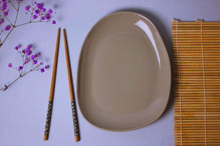 Table setting for sushi dinner. Gray empty oval plate, wooden chopsticks, bamboo sushi roll mat top view. Style tableware. Small purple Gypsophila flowers on a mauve blue table flatly. Asian Cuisineの写真素材