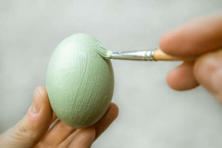 Hand holds brush and paints egg. A process of painting an Easter egg with light green paint. Paint brush in woman's hand on gray background. tradition and people concept. Spring season, April holiday.の写真素材