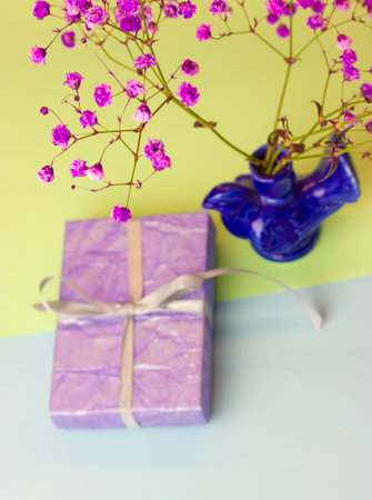 Purple gift box on light blue green festive background, blue ceramic bird shaped vase with purple flowers top view. Birthday greetings, Mother's Day congratulations. vertical wallpaper. selective focusの写真素材