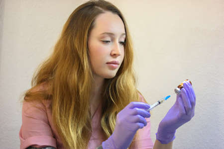 Young white woman nurse in pink robe purple gloves is taking hyaluronic acid filler into syringe for an injection. A cosmetic beautician doctor. Beauty Injection, plastic surgery, spa, care concept.の写真素材