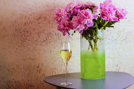 Bouquet of pink peonies in a green vase stands on a table in modern interior, a glass of champagne stands nearby. House warming, holiday at home concept. Spring, summer flowers in bloom. Place for textの写真素材