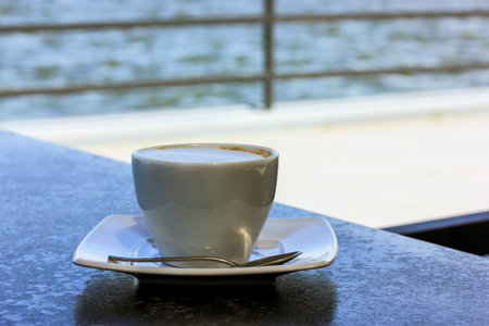 White coffee cup with cappuccino, latte, macacino stands on black table on cafe terrace on a sea, ocean shore in a sunny summer morning. Relaxing in the fresh air. Breakfast in a cafe, restaurant.の写真素材