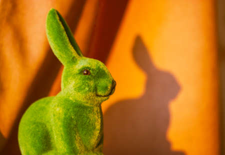 Green cute funny Easter bunny with long ears left shadow on orange brown background in springtime. Easter holiday in April. Toy decorative hare - a symbol of Easter - with copy space. Smiling rabbit.の写真素材