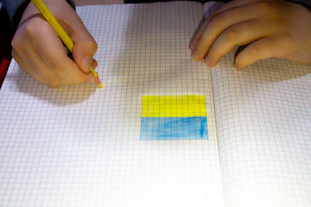 A child, kid draws in checkered notebook a yellow blue flag of Ukraine. Ukrainian boy is an artist. Children's hands and yellow pencil. A symbol of independence. Russian war in Ukraine 2022 concept.の写真素材
