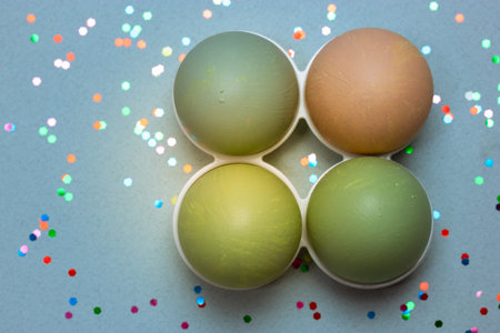 Four pastel-painted Easter eggs in egg tray stand on blue gray background with multicolored confetti...