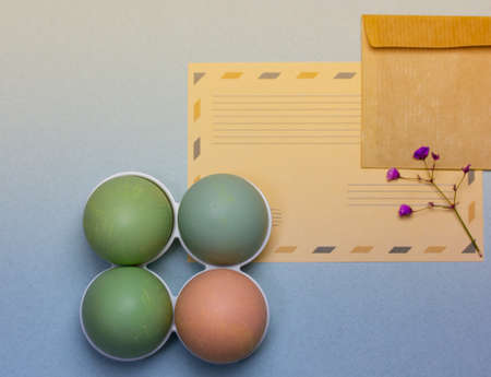 Easter card - brown letter envelope with space for text decorated with violet flowers. Four pastel colored Easter eggs on egg tray on blue gray background top view. Easter congratulations background.の写真素材