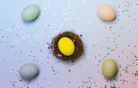 Easter composition with multicolored painted eggs lying on blue gray table top view.Yellow chicken egg in a nest in the center on blue-gray background with sequins. Celebration spring holiday in Aprilの写真素材