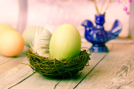 Green painted Easter egg in bird's nest with white feather on wooden table. Rustic Easter background. April holidays postcard. Easter egg wallpaper. Vase in the form of a blue bird on a background.の写真素材