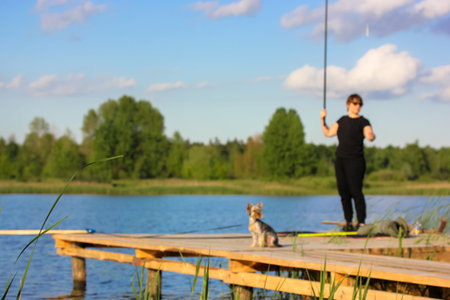 Woman with a fishing rod in hand and small Yorkshire Terrier dog standing on wooden bridge, pier, footbridge on a river, lake at summer day. Nature recreation. Beautiful landscape. Blurred photographyの写真素材