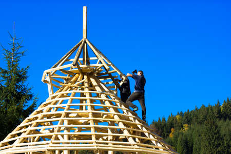 Bukovel, Ukraine. October 1, 2021. Construction a roof with wooden beams. Builder man with an ax hammering nails, reconstructing a dome against blue sky. Building a house in a pine coniferous forest.のeditorial素材