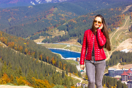 A happy young woman in a red jacket stands on mountain top and talks on phone. A traveler, tourist, adventurer in the mountains resort. Social media, mobile communication, mobile coverage concept.の写真素材