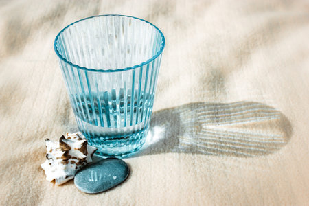Faceted blue glass standing on white sand next to a seashell. Refreshing drink still life with...