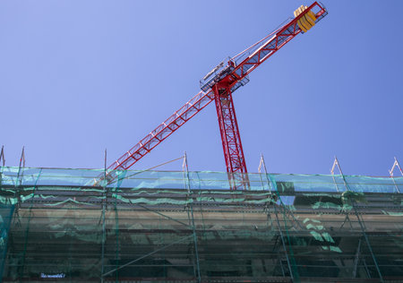 Red construction crane against blue sky at construction site. A facade of an old building closed for renovation, repair with green protective construction mesh. Engineering architectural business.の写真素材