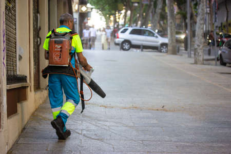 Madrid, Spain. June 1, 2022. City street cleaner. Utility worker in uniform cleans sidewalk of dust, leaves, debris with garden vacuum cleaner, vacuum blower. Cleaner man in urban scene.のeditorial素材