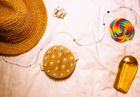Straw hat, anti-cellulite massager, bright sweet multicolored lollipop, sunscreen oil lie on a white sheet top view. Cosmetics for summer, beach season. female lifestyle concept. Women Accessorizes.の写真素材