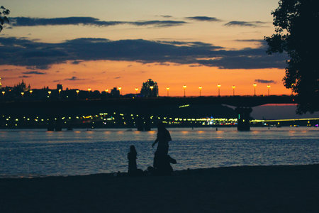 Night city panorama, calm water in a Dnipro river. City lights shining on a horizon. Silhouettes of people relaxing on a shore bay in cloudy twilights, evening, nighttime illumination. Kyiv, Ukraine.の写真素材