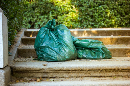 Green plastic trash bags lying on the steps in the park on a green natural blurred background. Cleaning environment. Pollution, trash collection, gardening on a backyard. Rubbish, waste bag.の写真素材