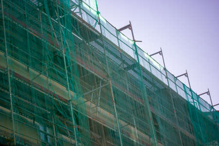 Multistory building facade covered with green protective construction mesh on blue sky background. Construction, reconstruction of residential buildings, house renovation, skyscrapers in big city.の写真素材