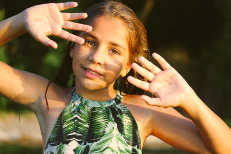 Beautiful little teen girl in a green swimsuit posing standing in water of river, lake, sea in summer. A cute child covers her face from the sun in summer. Female kid on tropical resort in summertime.の写真素材