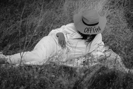 Black and white photograph of a girl, a young woman in a straw hat with text Offline lying on the grass. The enigmatic lady is resting on a picnic, hiding her face. Faceless people outdoors on nature.の写真素材