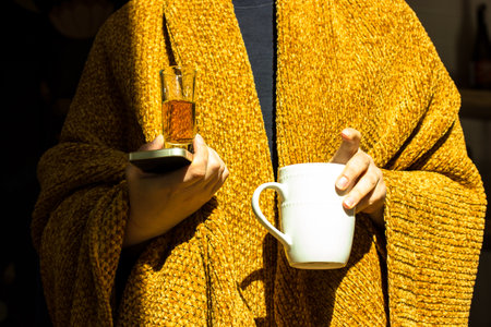 Faceless person wrapped in warm brown-yellow mustard rug, blanket holds in hands white tea, coffee cup, shot glass with herbal tincture, whiskey, brandy alcoholic beverage warming up in autumn, winterの写真素材