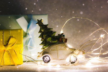 Toy retro car with Christmas tree on roof, blue and yellow gift boxes among led garland lights on dark silver background. New Year's Eve 2023. Winter sales, shopping concept. Winter holidays magic.の写真素材