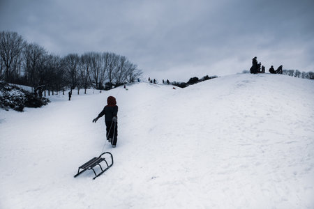 A little boy in a dark jumpsuit pulls a sled behind him as he climbs a snow-covered slope to sled down. Children's outdoor activities fun in a city park on winter vacation. First snow in cold season.の写真素材