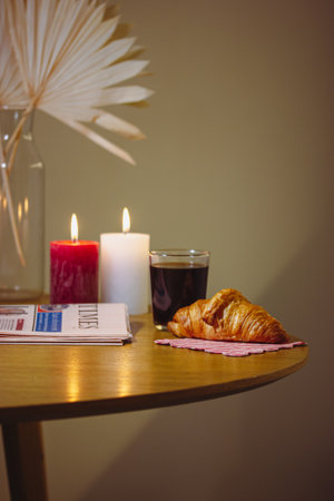 Vertical still life with tea, coffee glass, red white burning candles on wooden kitchen table. Croissant for breakfast in cozy home. Warm lighting indoors Cozy scene food and drink. Morning newspapersの写真素材