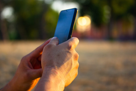 Cell phone in strong man's hands. Faceless male, guy uses smartphone gadget sitting in park in sunny...