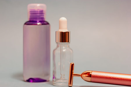 Vibrating golden facial massager, bottle with serum, lavender massage oil dropper on grey background Female body skin care. Luxury beauty tool. Prolonging youthfulness. Pulse face massaging instrumentの写真素材