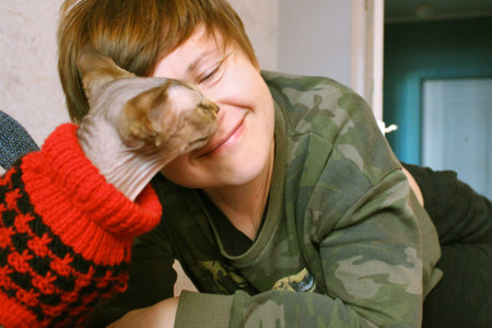 A happy smiling woman plays with Canadian Sphynx cat in a red jumper at home. Contact, interaction, play with a beloved pet. Hairless kitty feline animal. Sharing love with animals. Sphinx fashion.の写真素材